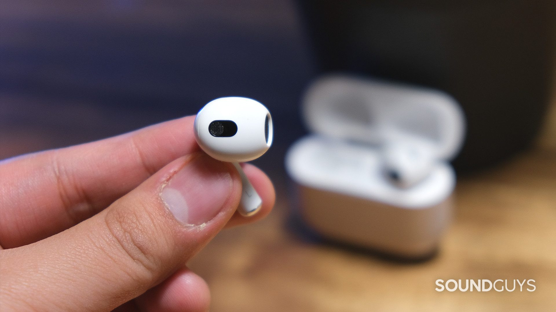Apple AirPods 3rd Generation_8 A hand holds a Apple AirPods (3rd generation) earbud by the stem to reveal the open-type fit and embedded sensors with the open case in the background.
