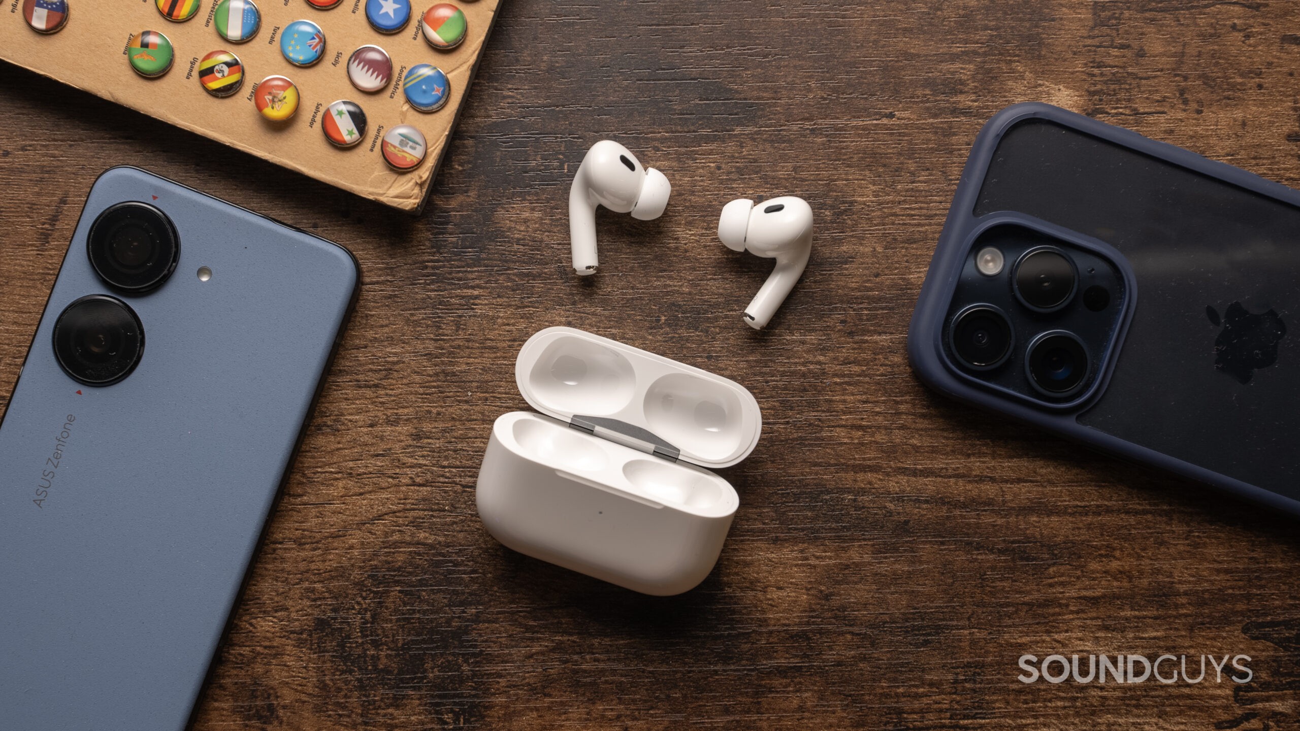 AirPods-Pro-2nd-Generation-USB-C-open-on-table Apple AirPods Pro (2nd generation) resting on a dark wood tabletop with the lid open and buds on display surrounded by phones.