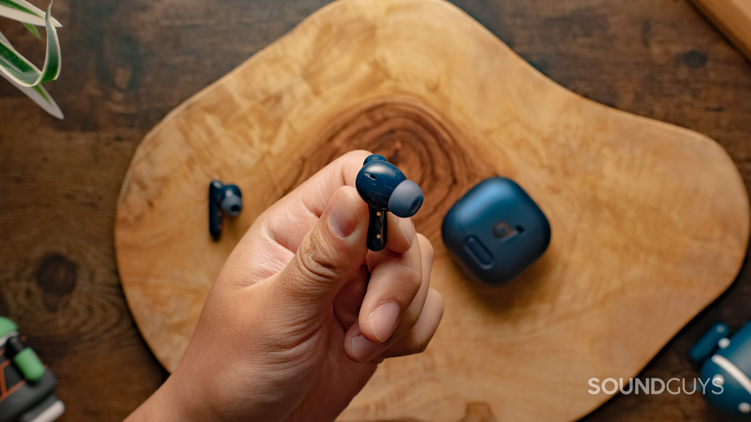Anker-Soundcore-Liberty-4NC-Bud-Image-#1 A hand holds one of the Anker Soundcore Liberty 4 NC by the stem with the case on a wood surface in the background.
