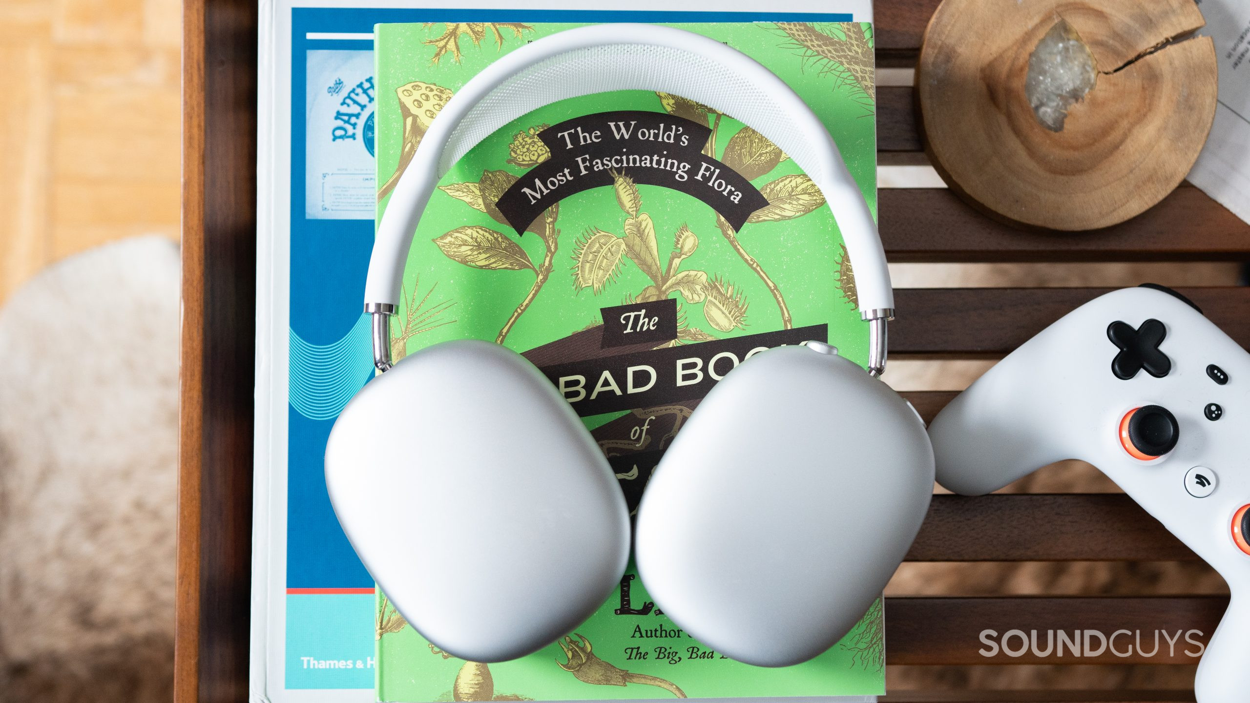 Apple AirPods Max rotating ear cups The white Apple AirPods Max lying flat on a green book with a white Google Stadia controller next to it.