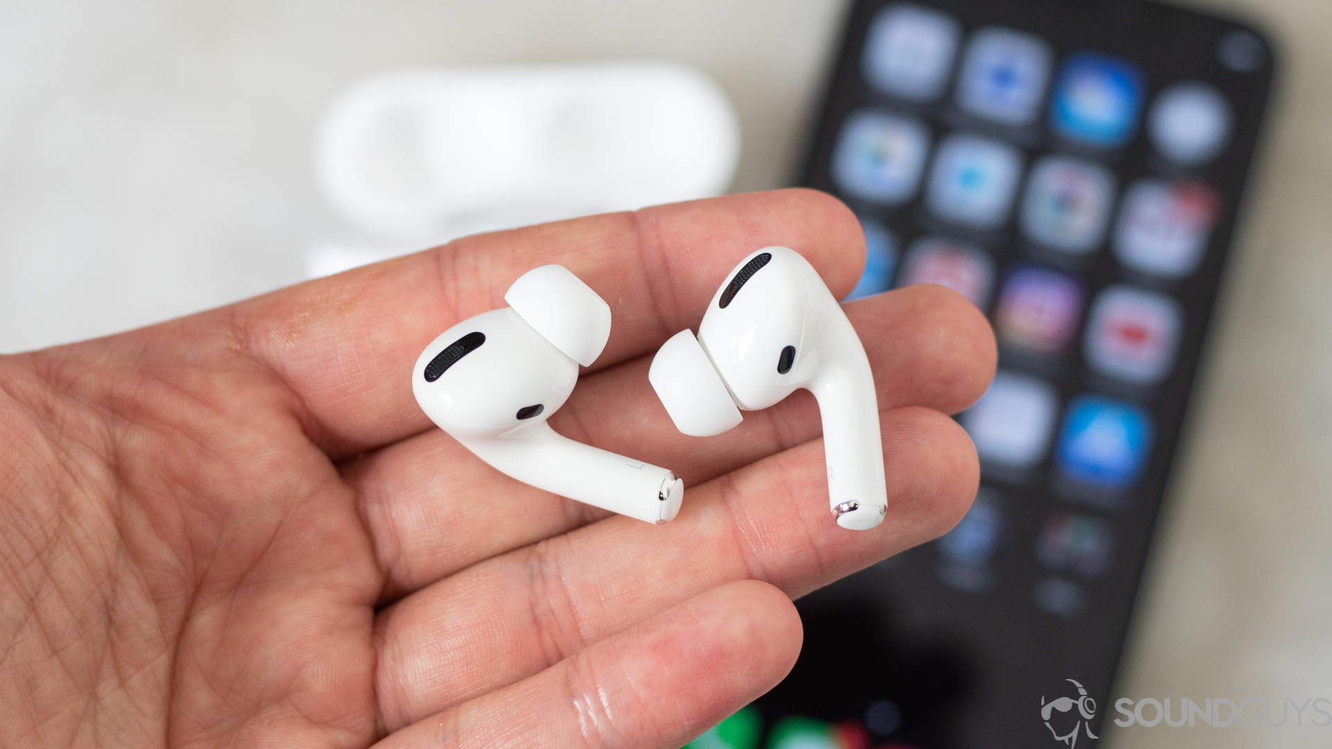 Apple AirPods Pro-11 The Apple AirPods Pro in a man's left hand (foreground) with an iPhone and the AirPods Pro wireless charging case in the background.