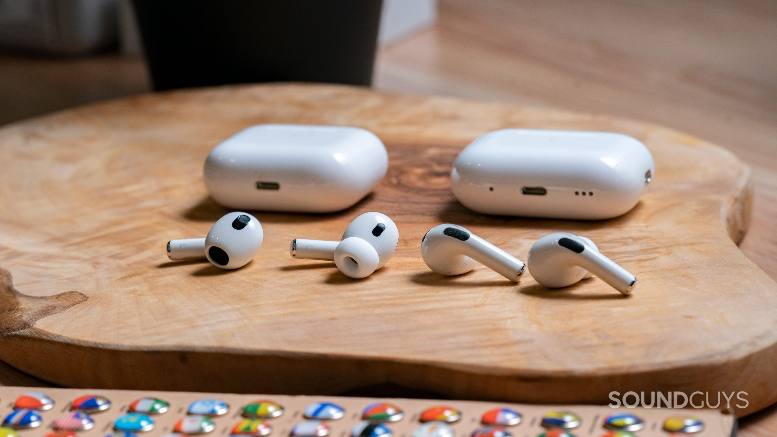 Apple Airpods Pro Gen 2 vs. Apple Airpods Gen 3 Buds Comparison The Apple AirPods Pro Gen 2 and the Apple Airpods Gen 3 sitting on a wooden surface with their cases behind them.