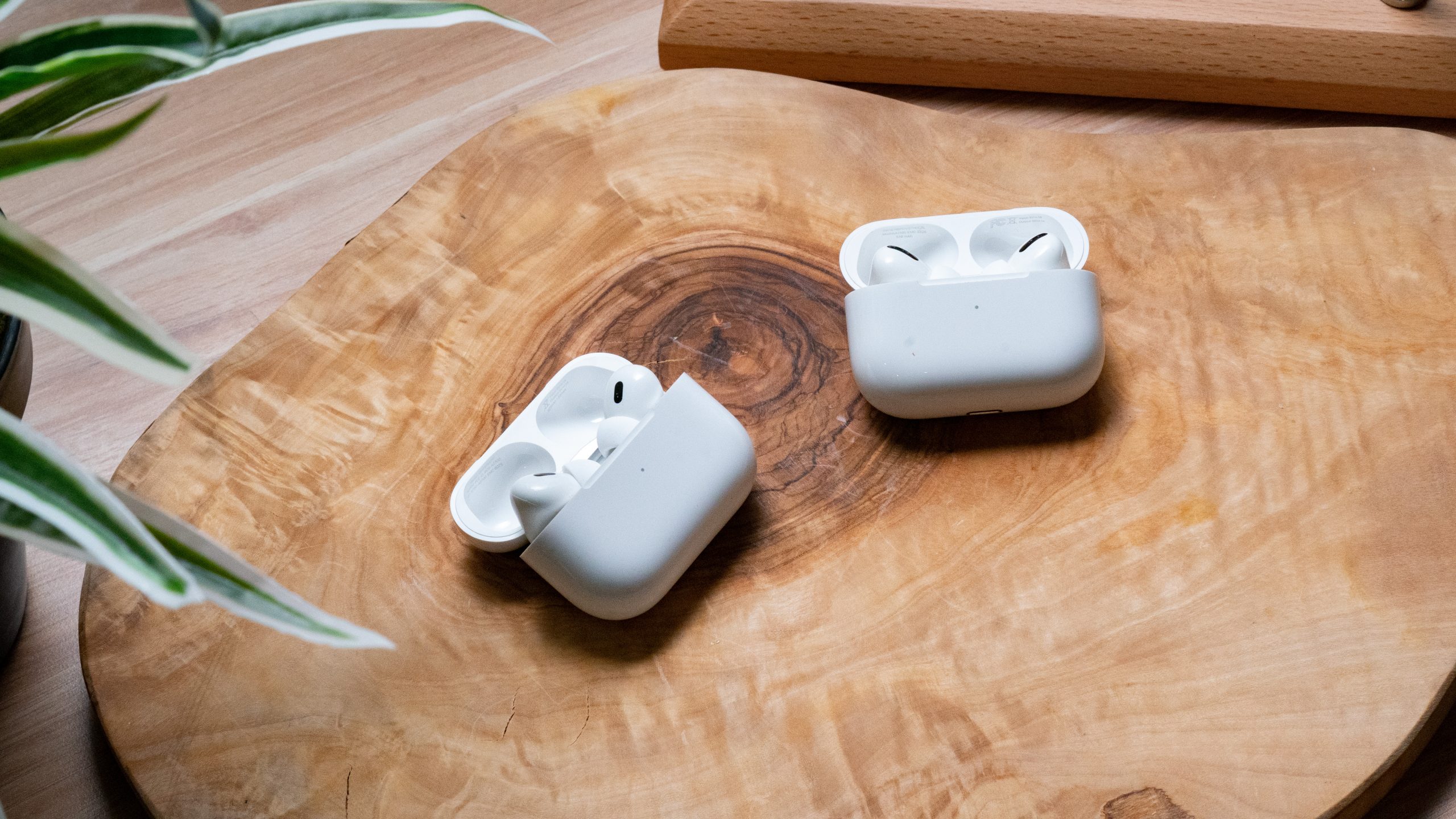 Apple Airpods Pro Gen 2 vs. Apple Airpods Pro Gen 1 Hero Image #2 The Apple AirPods Pro (2nd generation) lays on a wooden surface next to the Apple AirPods Pro (1st generation), earbuds in open cases.