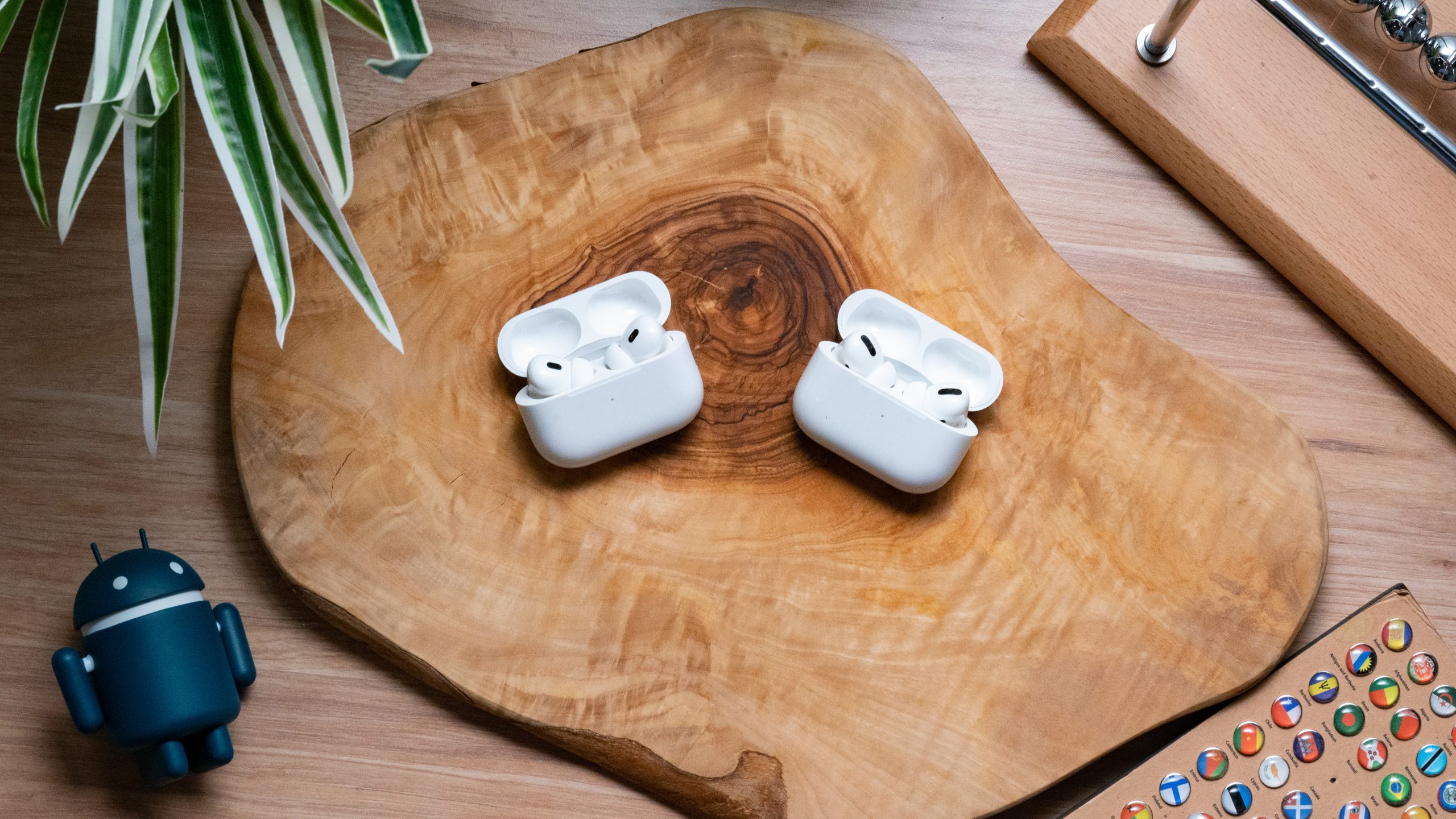 Apple Airpods Pro Gen 2 vs. Apple Airpods Pro Gen 1 Hero Image The Apple AirPods Pro (2nd generation) lays on a wooden surface next to the Apple AirPods Pro (1st generation), earbuds in open cases, shot from above.