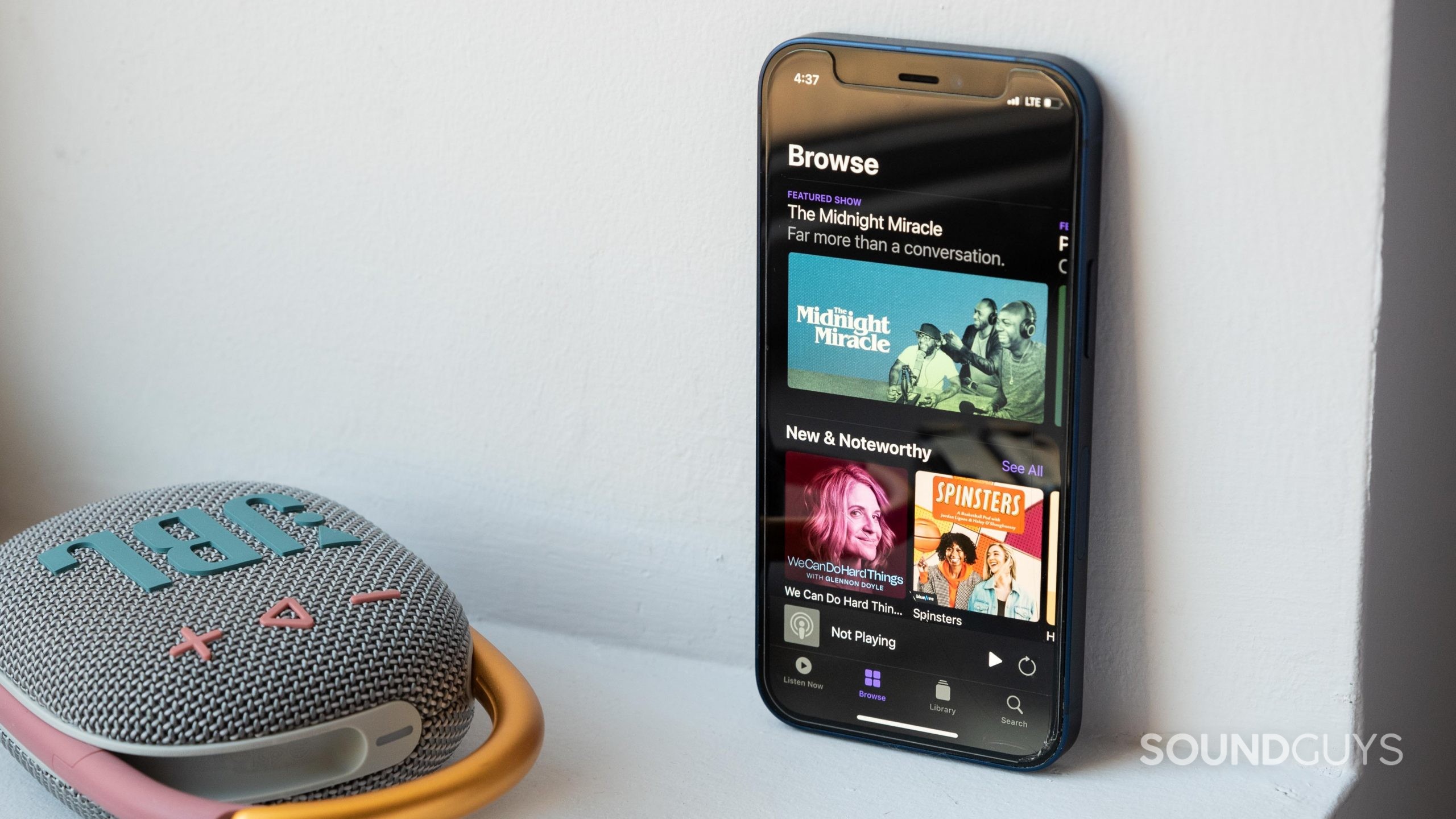 Apple Podcasts review mobile app-1 An iPhone 12 Mini rests against a wall, next to the JBL Clip 4 Bluetooth speaker, and the phone displays the Apple Podcast app homepage.