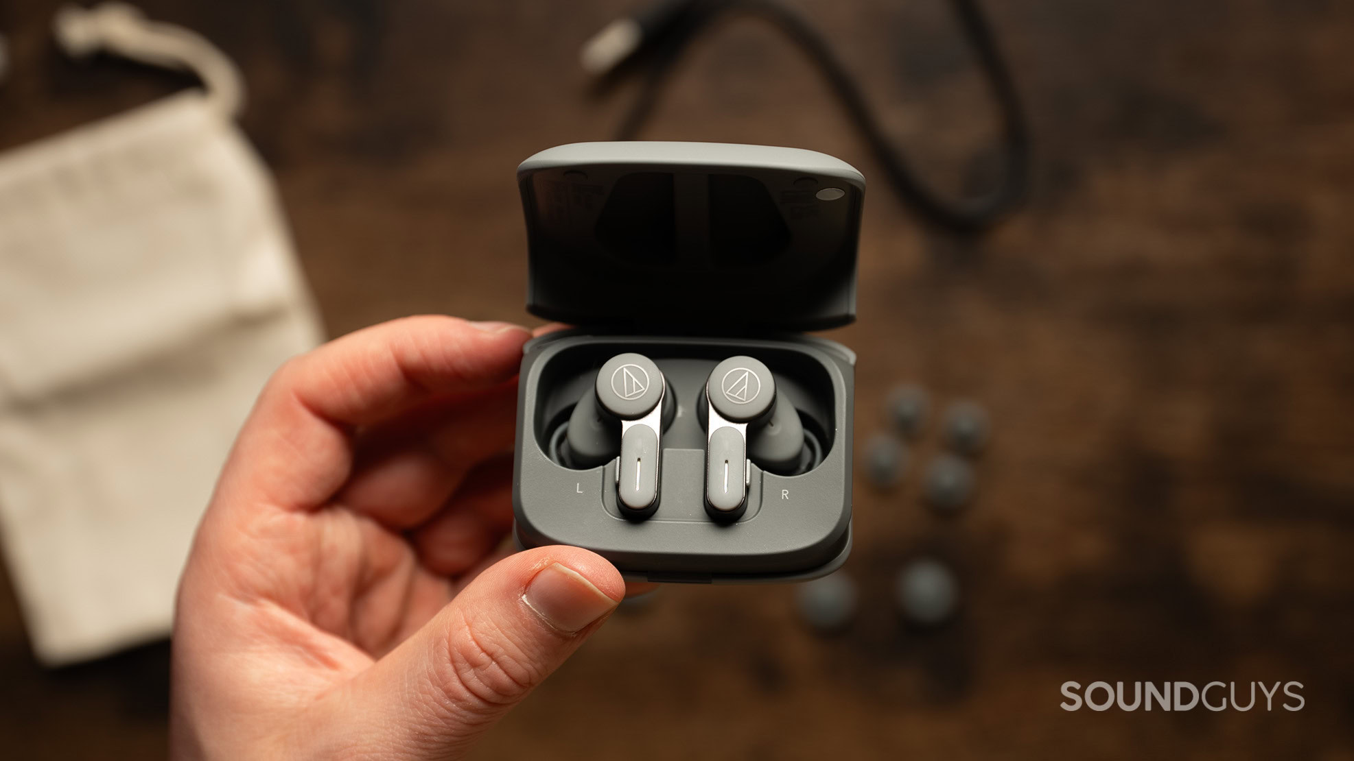 Audio Technica ATH-TWX7 hero 2 A hand holds the Audio-Technica ATH-TWX7 earbuds inside their charging case over a wood table.