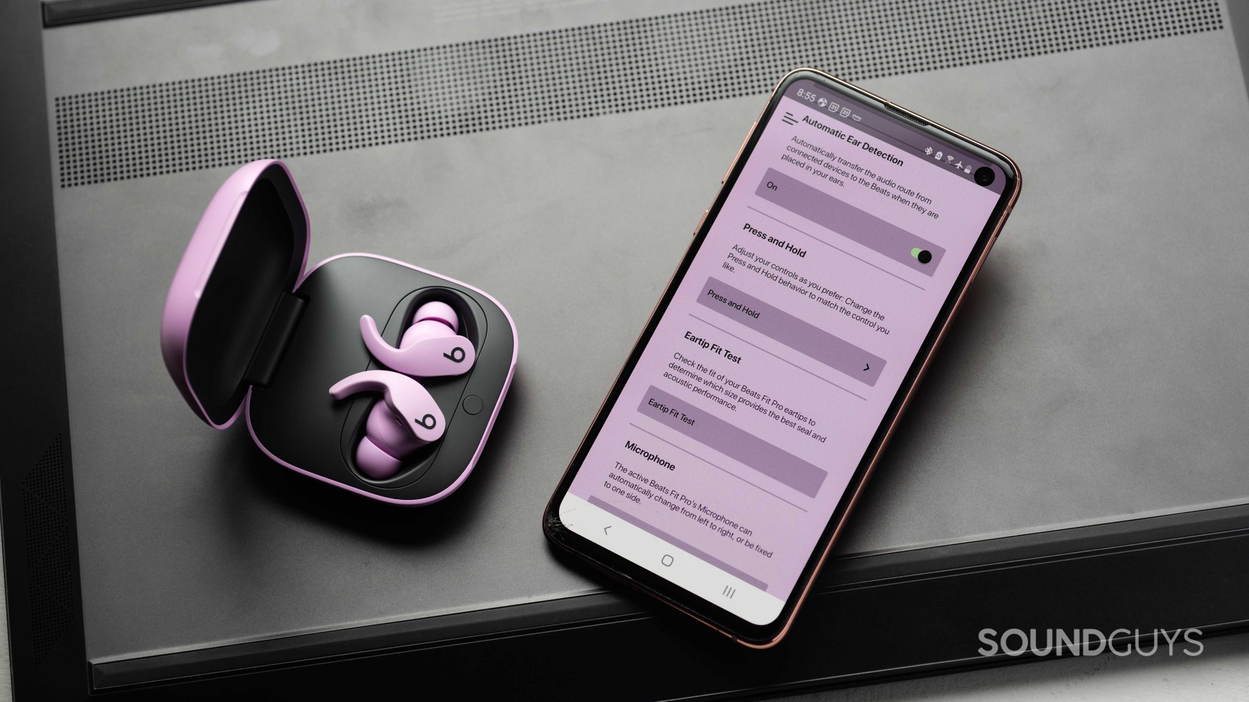 Beats Fit Pro noise canceling true wireless earbuds mobile app The Beats Fit Pro noise canceling true wireless earbuds in the open charging case and next to a Samsung Galaxy S10e with the Beats app open. The app has a purple tint to it, presumably to match the earphones.