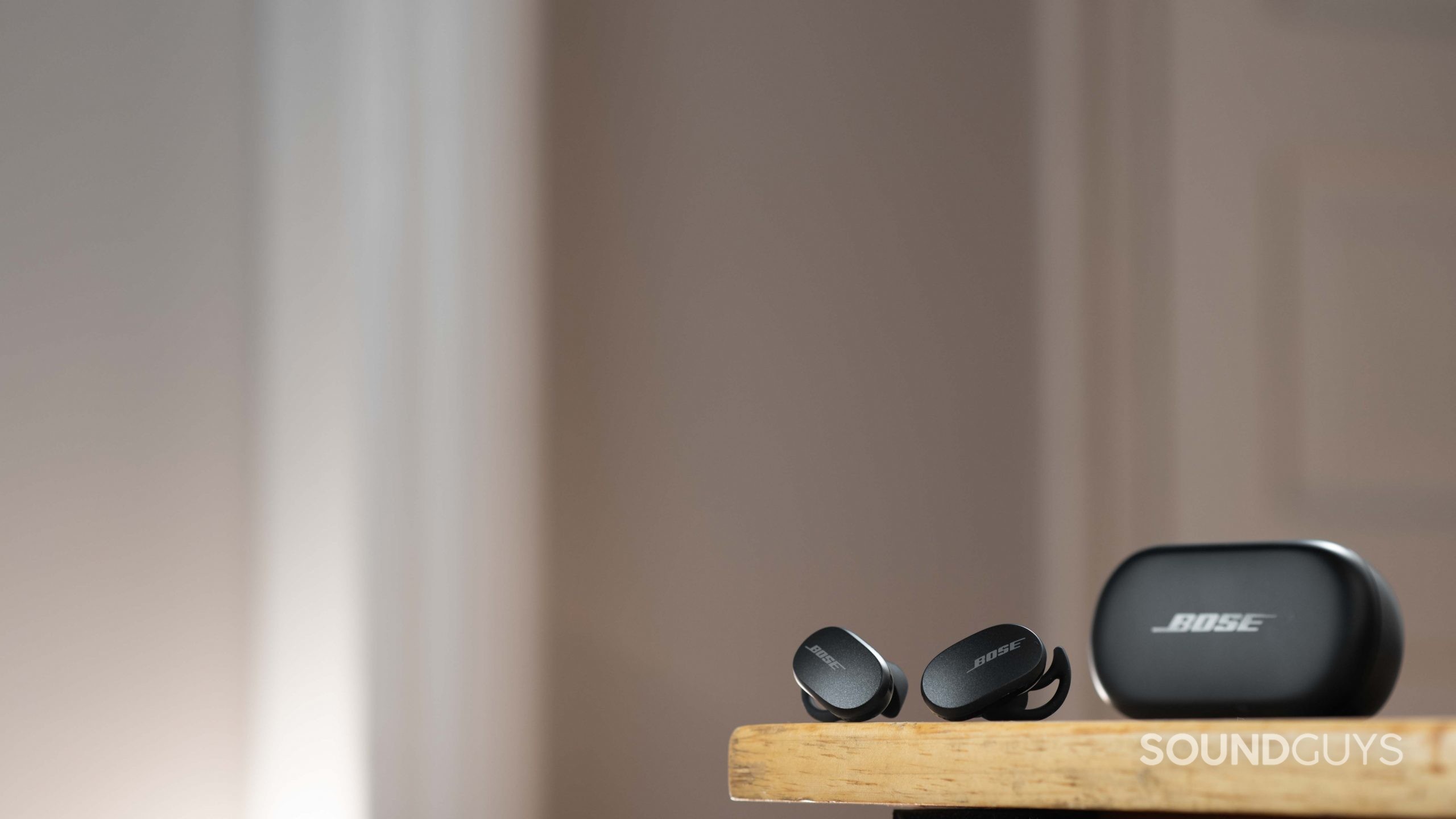 Bose QuietComfort Earbuds noise canceling true wireless-3 The Bose QuietComfort Earbuds noise canceling true wireless earbuds rest next to the closed charging case on a wood surface.