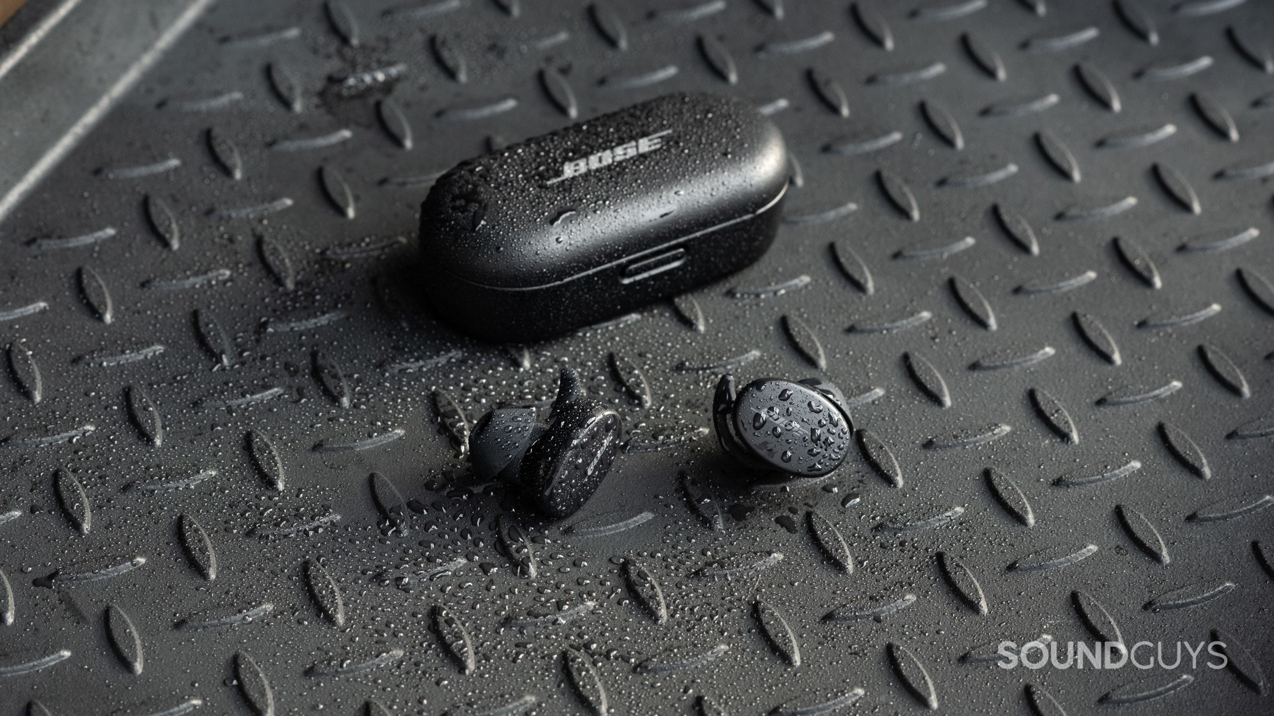 Bose Sport earbuds-10 The Bose Sport Earbuds true wireless workout earbuds sit outside of the closed charging case, all objects are covered in sprinkles of water.