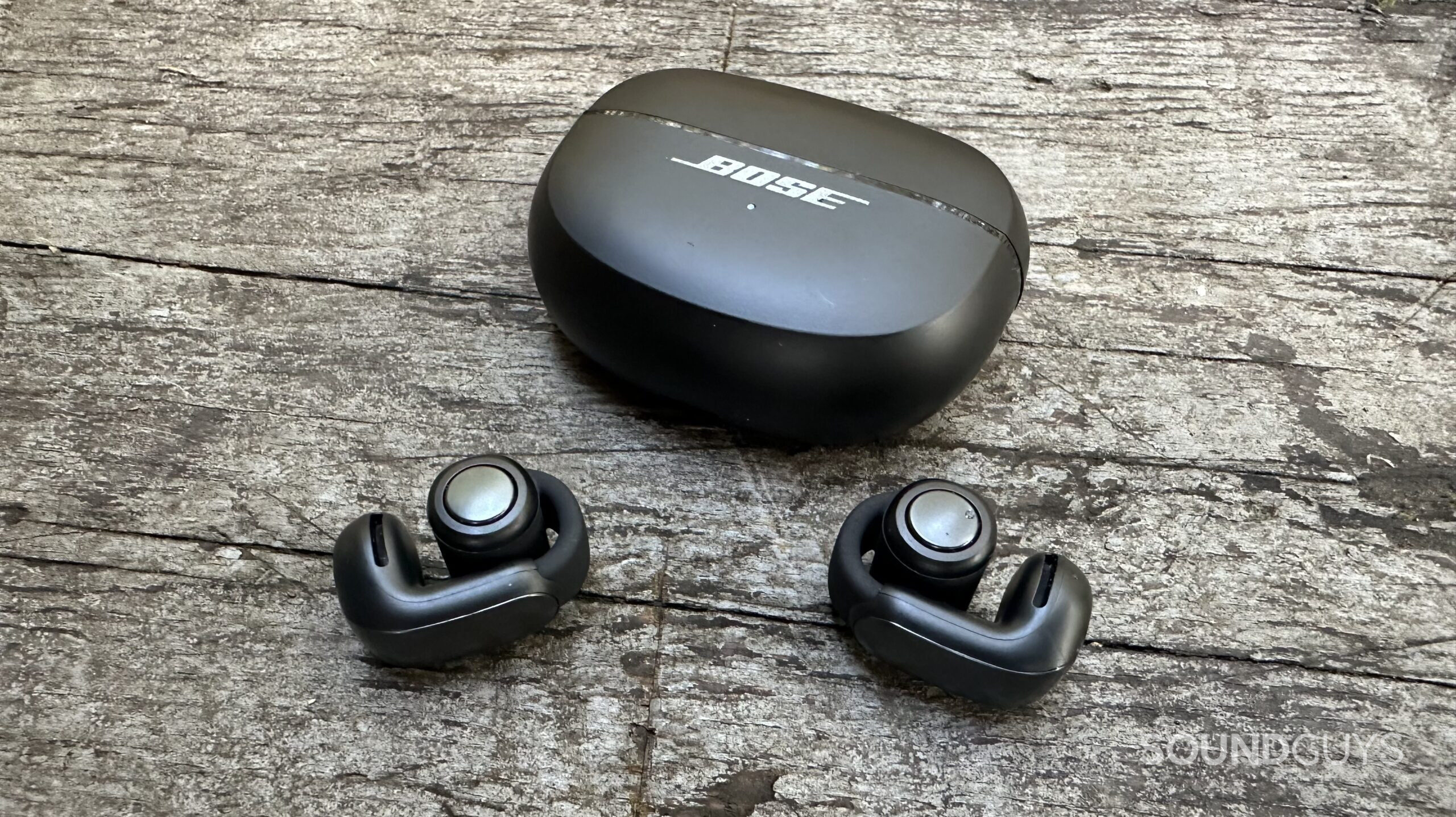 Bose UOE Closed case and clips Bose Ultimate Open Earbuds and their charging case on a grey bench