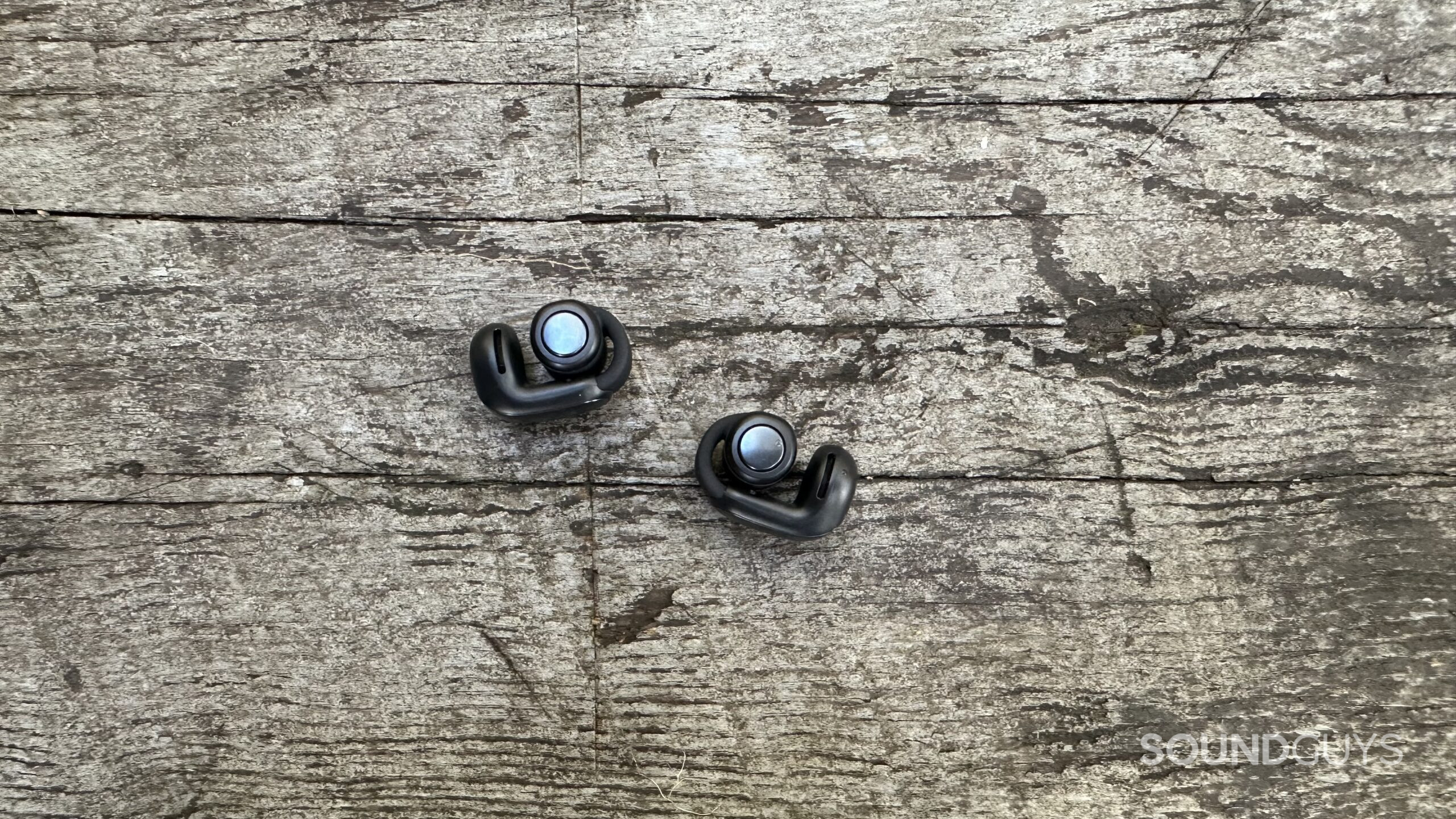 Bose UOE control buttons A pair of Bose Ultimate Open Earbuds with control buttons facing up on a grey bench
