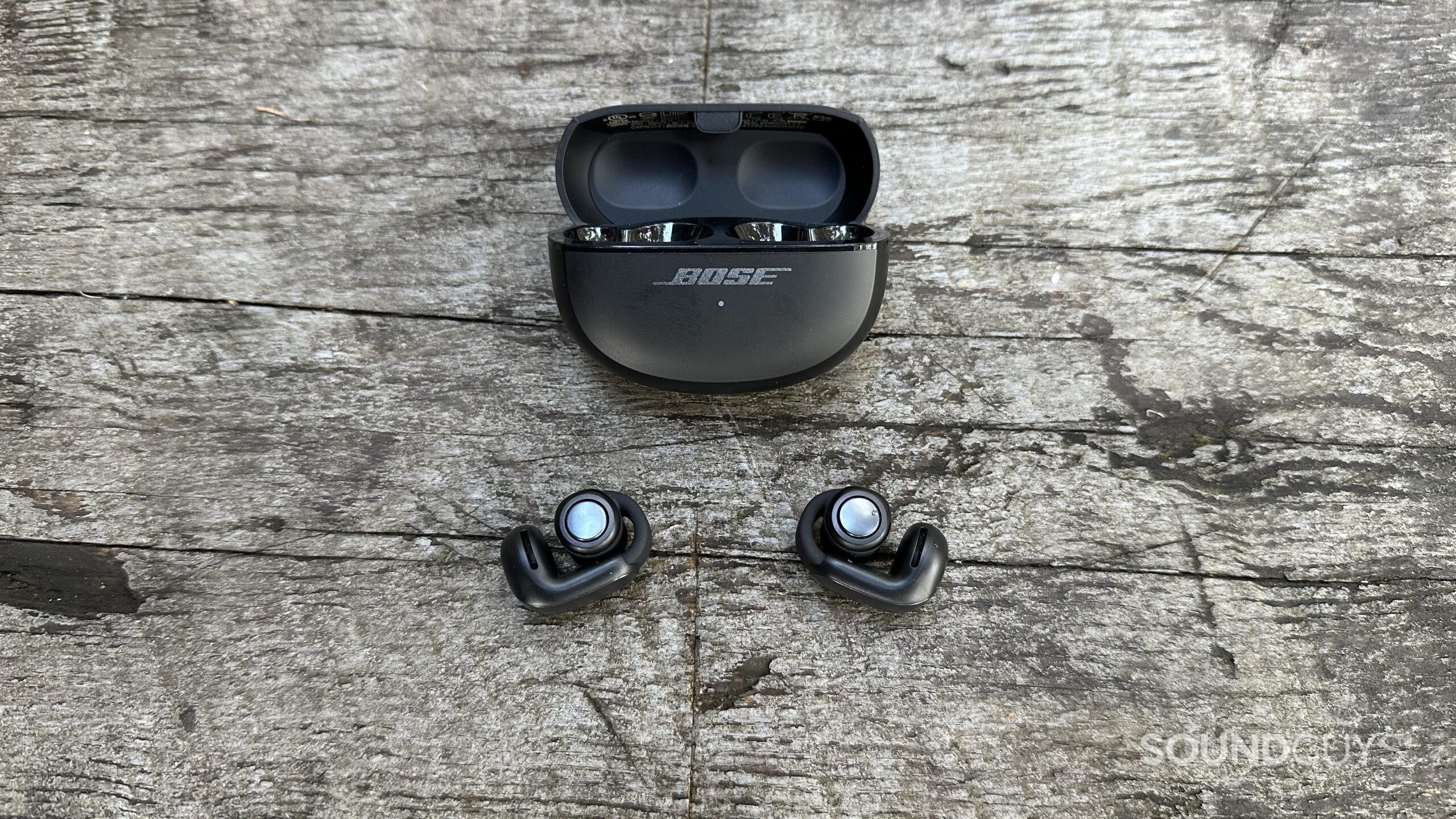Bose UOE Open case and clips 2 A pair of Bose Ultimate Open Earbuds and open charging case sitting on a grey bench