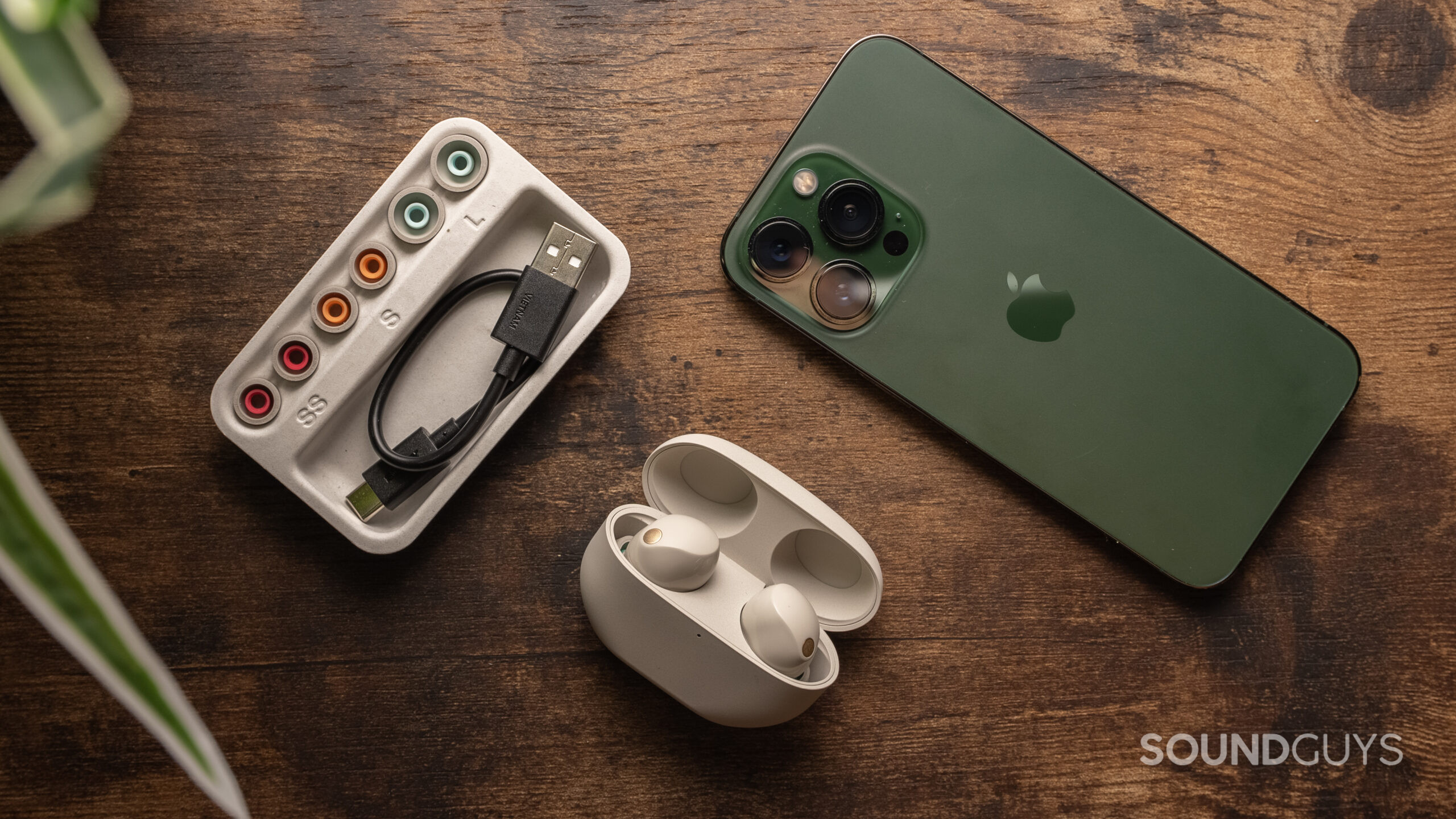 Sony WF-1000XM5 with accessories The Sony WF-1000XM5 charging case open with the earbuds inside beside a green iPhone 13 Pro and the ear tip selection and USB charging cable