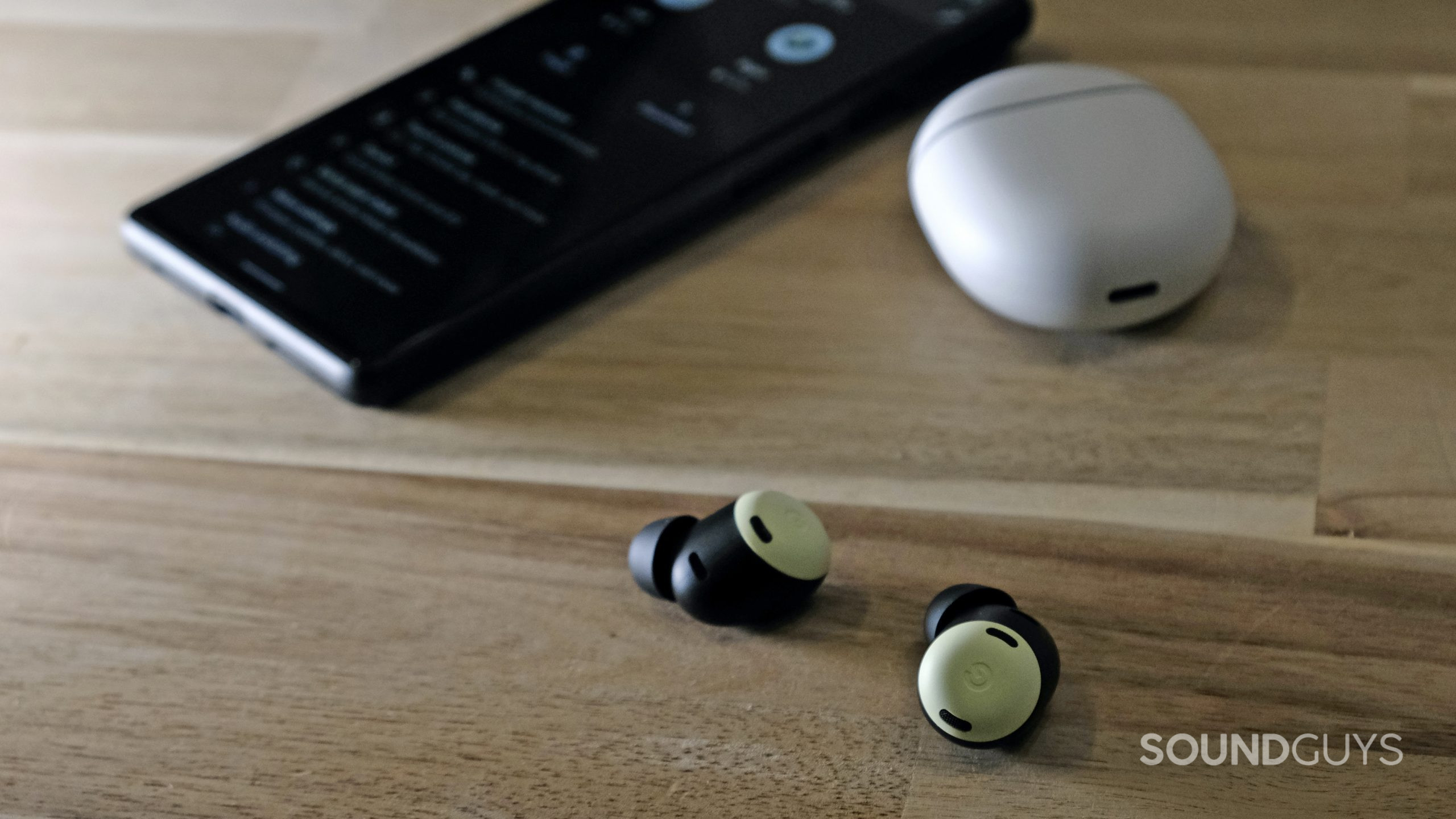 Google-Pixel-Buds-Pro-buds-on-table The Google Pixel Buds Pro lays on a wooden table with a Google Pixel 6 behind it.