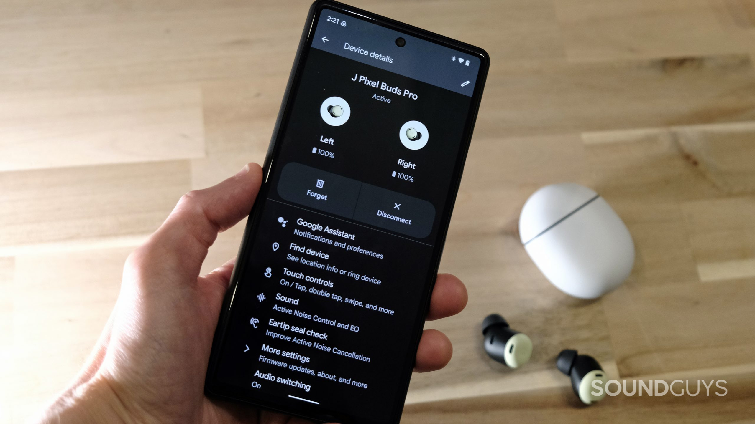 Google-Pixel-Buds-Pro-Pixel-settings-app A hand holds a Google Pixel 6 displaying the Google Pixel Buds Pro controls in the Settings app, with the true wireless earbuds on a wooden table in the background.