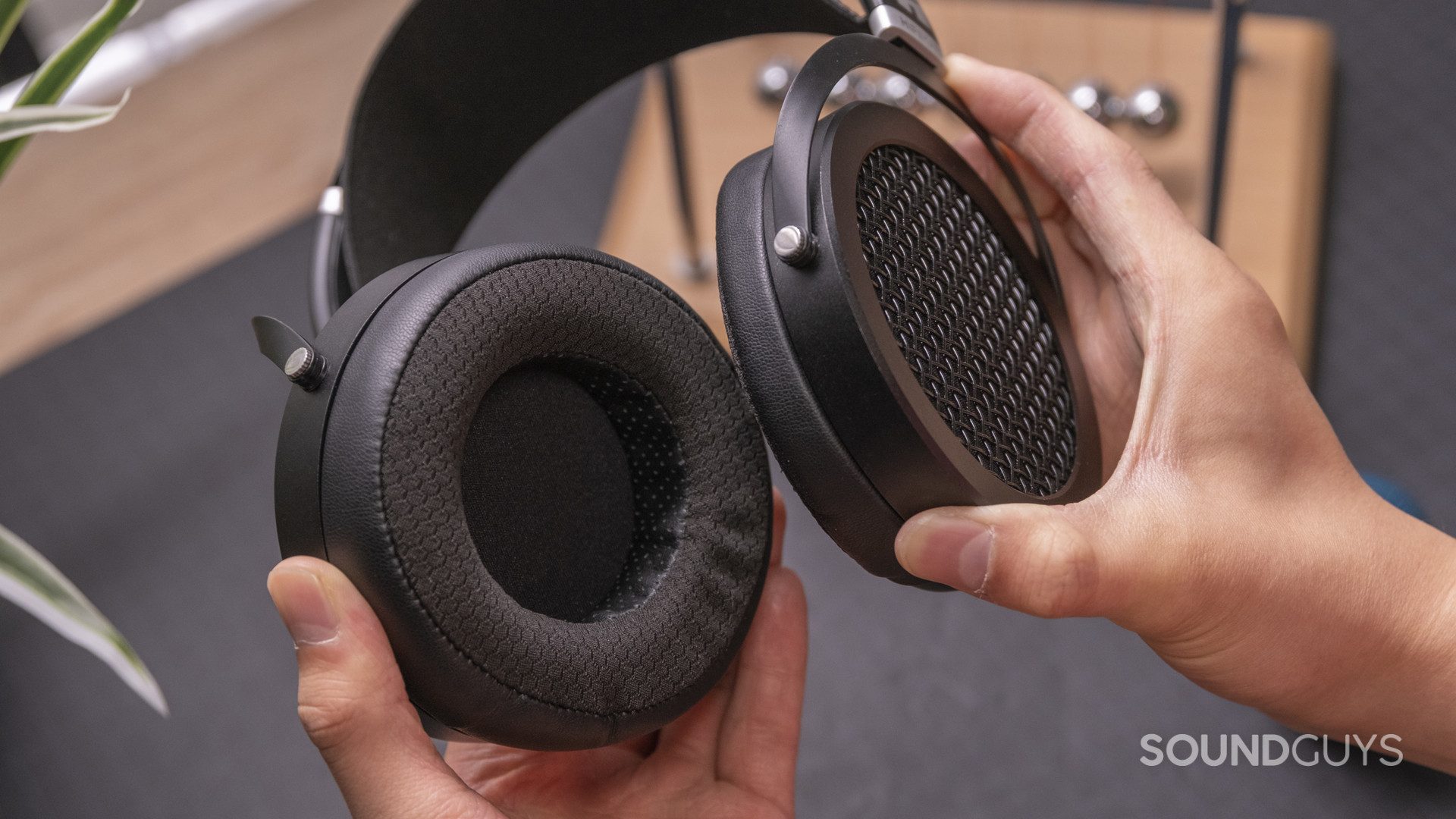 hifiman-sundara-pads-direct Hands hold the HiFiMan Sundara open-back headphones in front of a black backdrop with wooden accessories.
