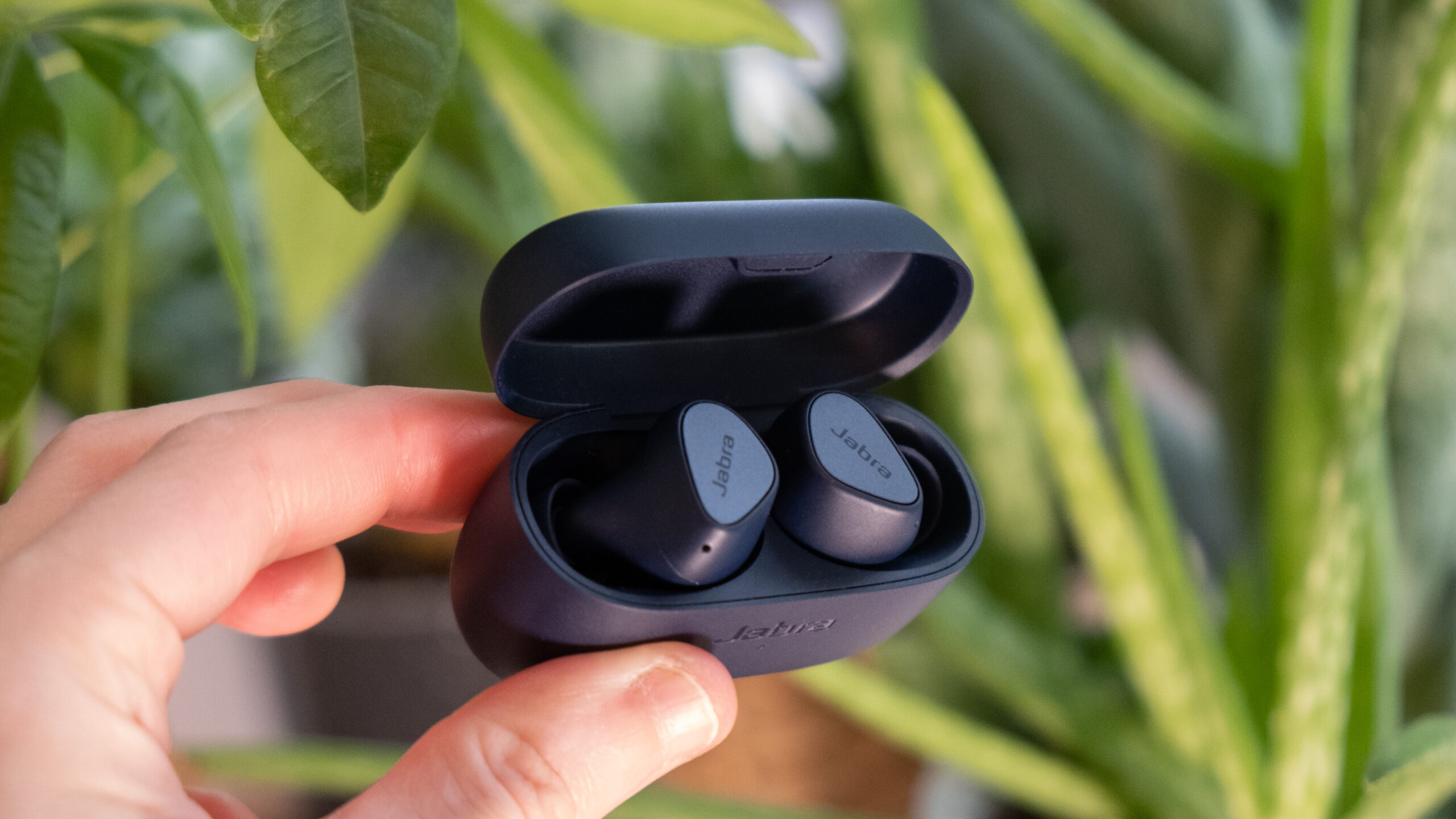 jabra-elite-4-in-hand-center A hand holds the Jabra Elite 4 open in front of green foliage.