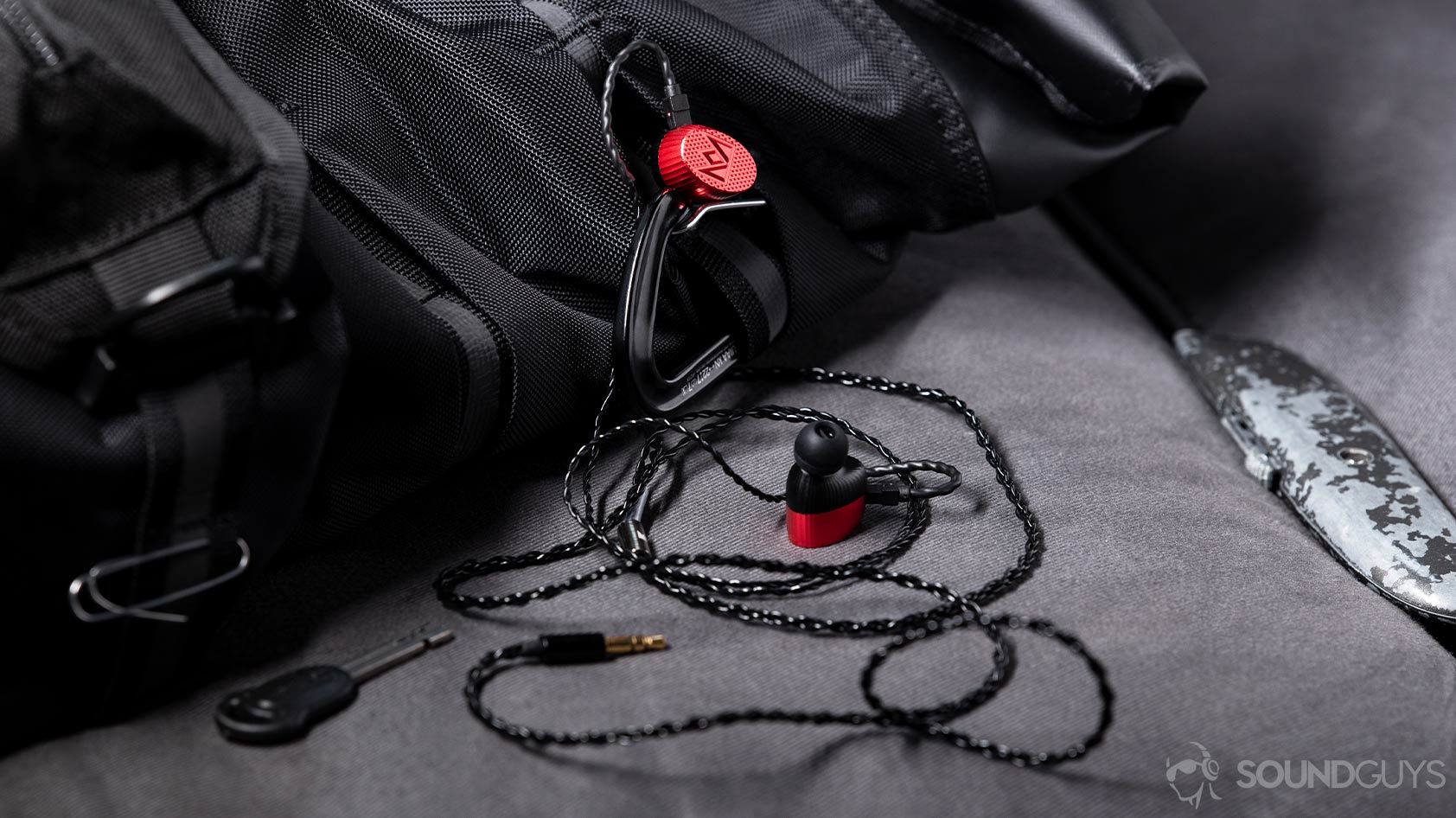 Massdrop x Noble Kaiser 10 Universal-in ear earbuds professional studio monitors backpack A level photo of the Massdrop x Noble Kaiser 10 Universal earbuds hanging off of a black back pack with a box cutter to the right side of the image.