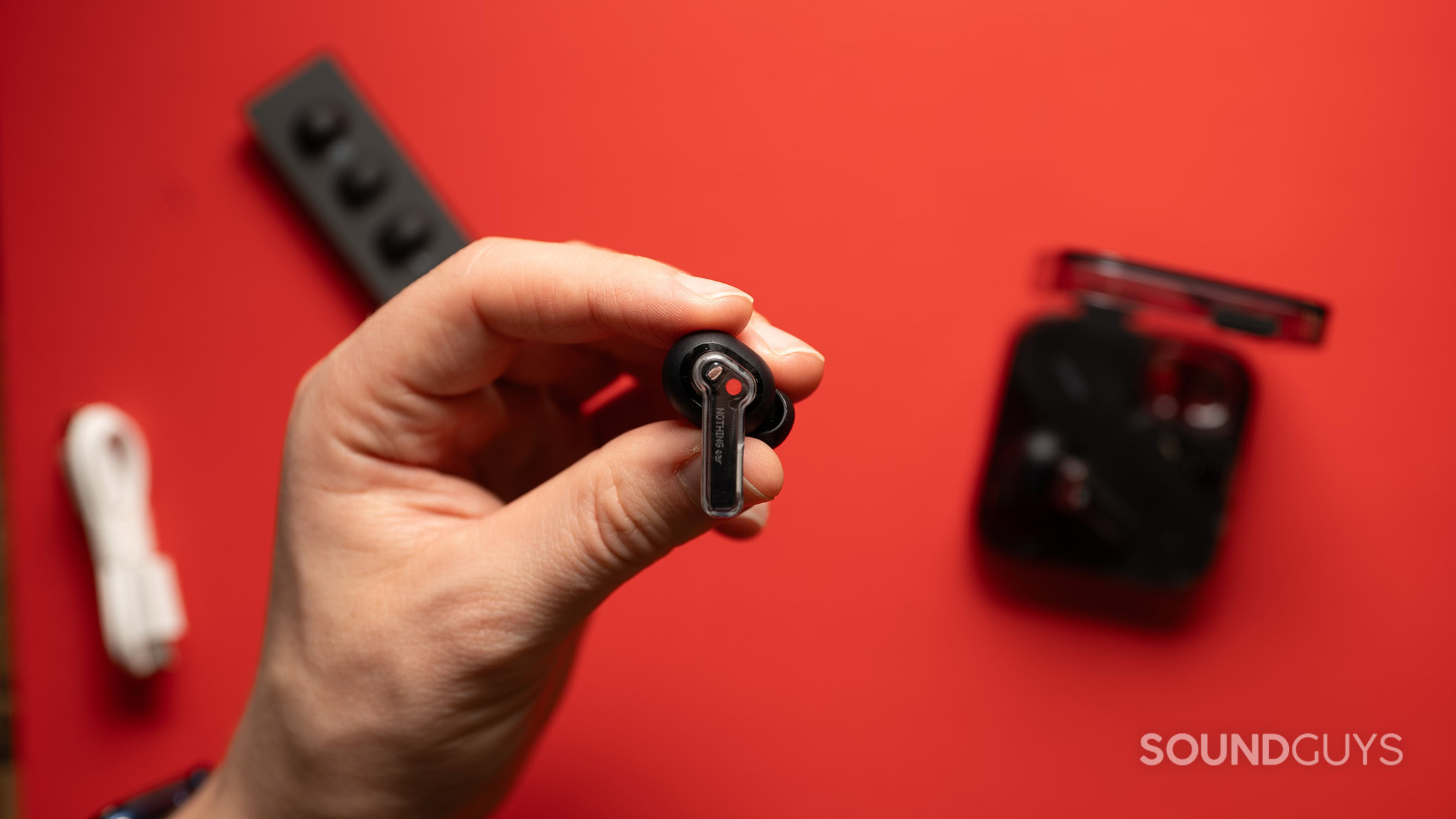 Nothing Ear earbud and accessories A hand holds a Nothing Ear earbud over a table with the charging case, extra ear tips, and charging cable.