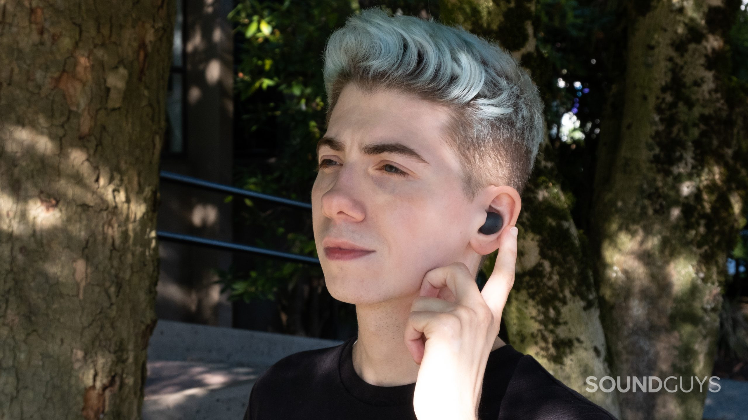 samsung-galaxy-buds-2-pro-in-ears A man wears the Samsung Galaxy Buds 2 Pro while facing left, and he's about to tap the touch pad with trees and concrete in the background.