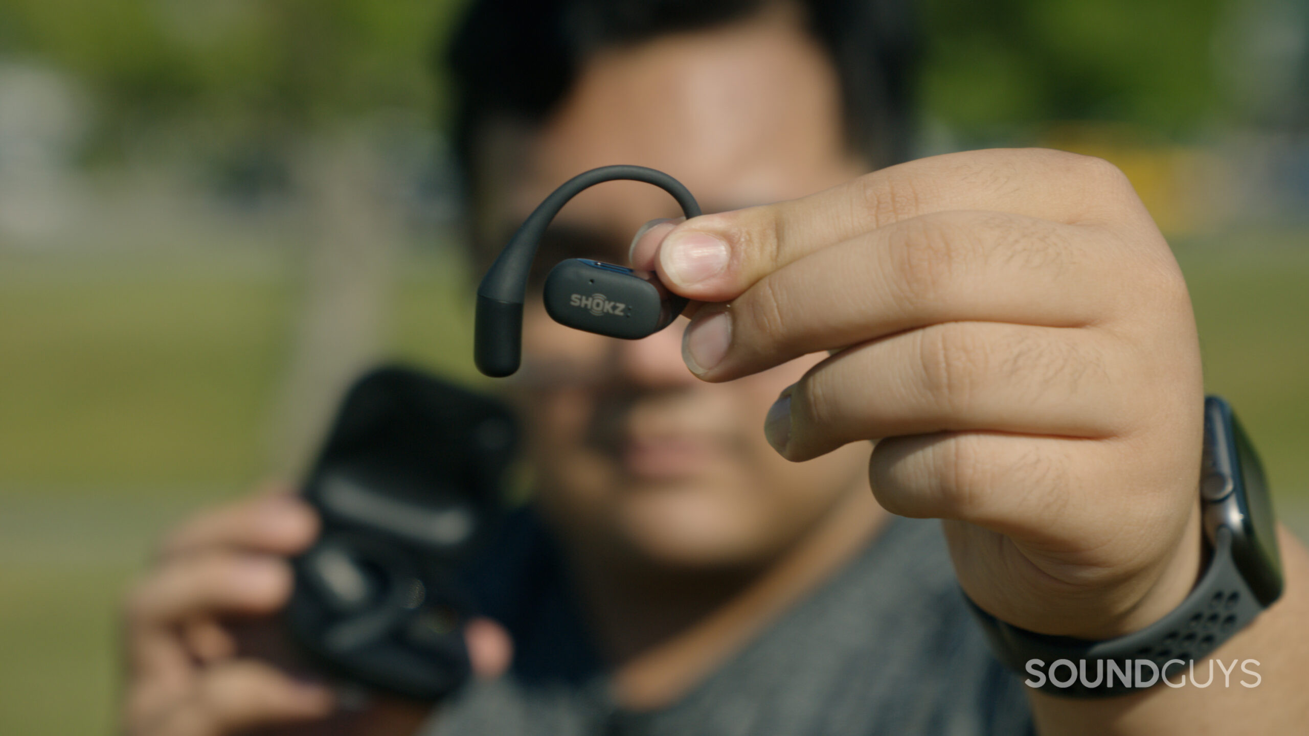 Shokz OpenFit in hand A man out of focus holds the Shokz OpenFit in focus.