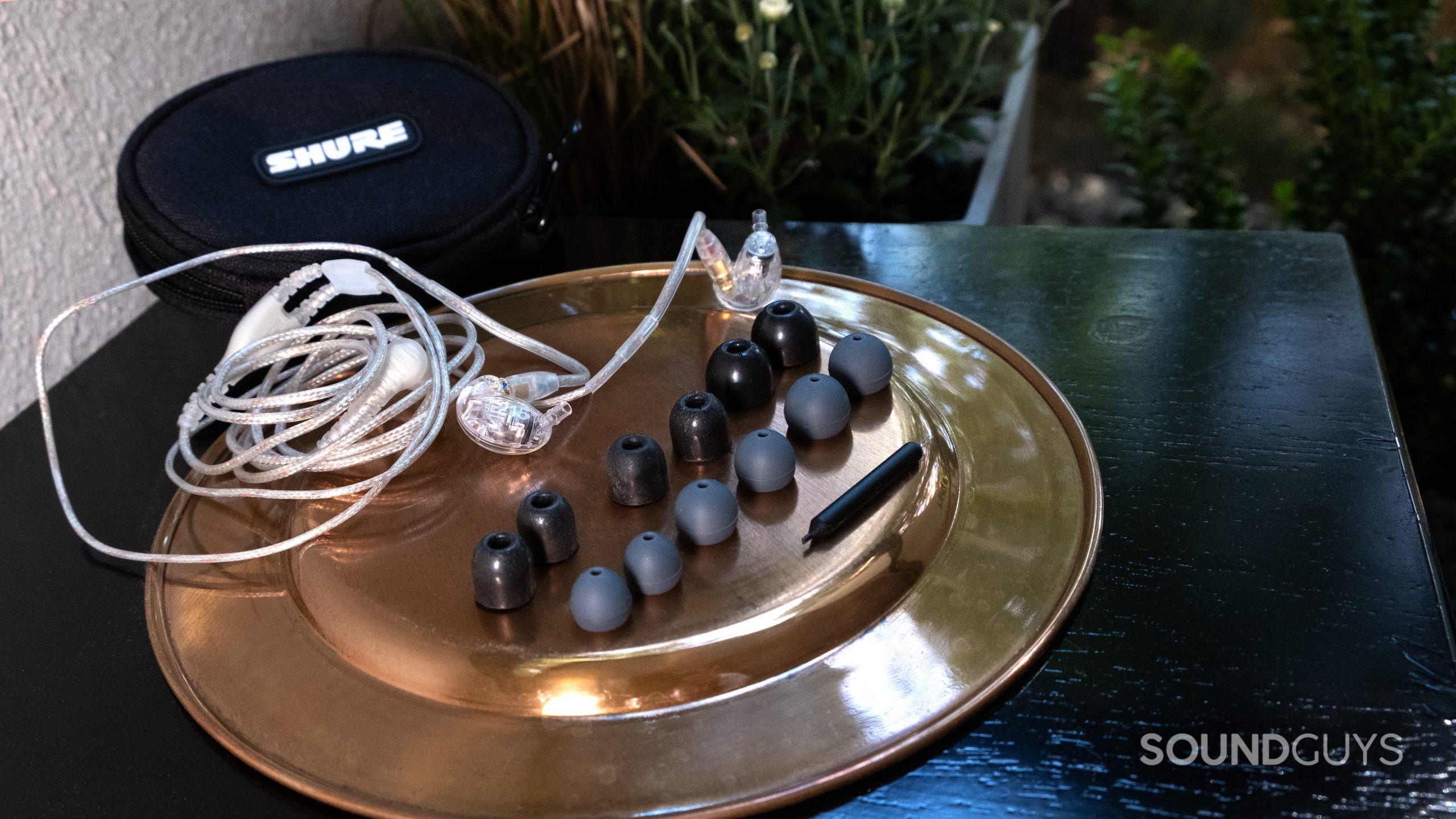 shure se215 accessories On a brass colored surface the full set of ear tips and the Shure SE215 sit outside on a black table with plants in the background.