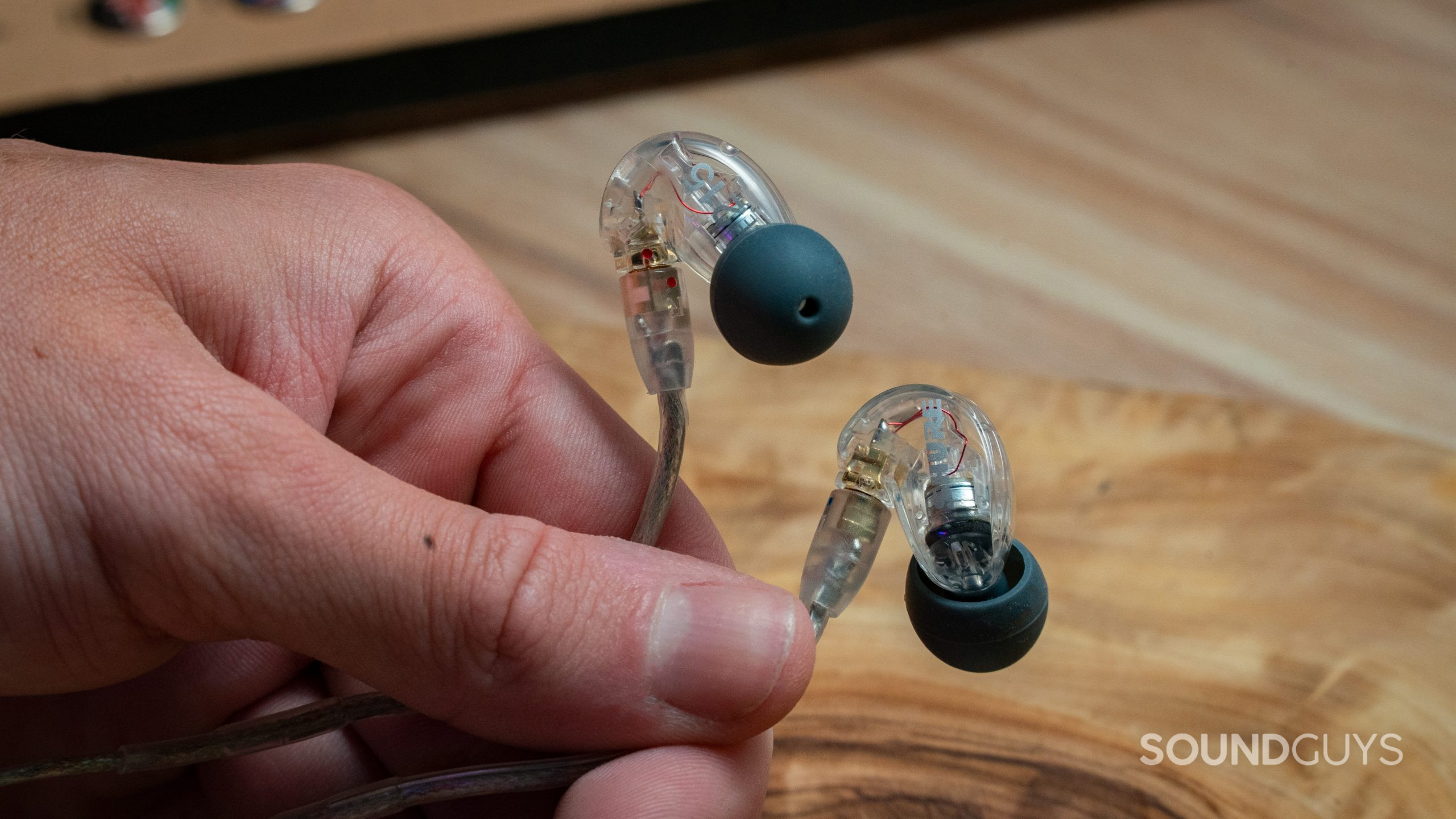 SHURE_SE215 IEMs handheld A hand holds the Shure SE215 in front of a wood table.