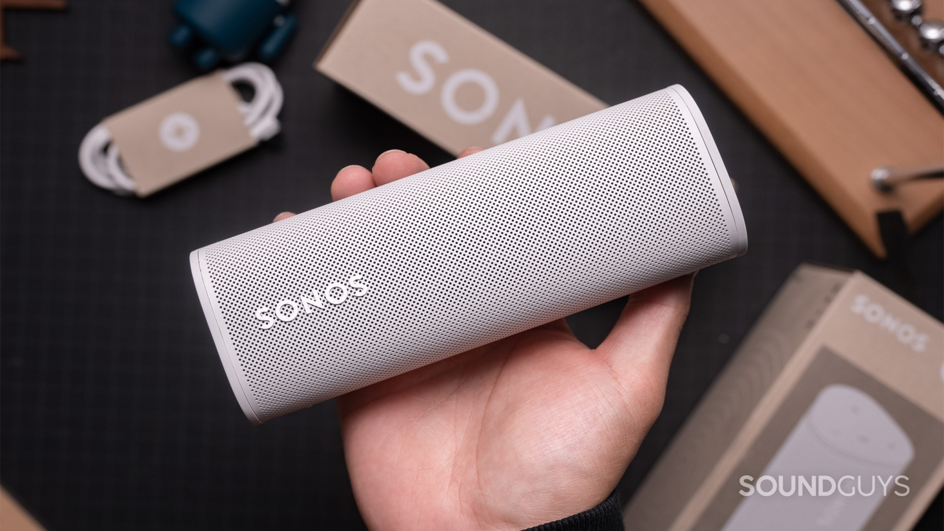 Sonos Roam Bluetooth smart speaker hand holds accessories A hand holding a white Sonos Roam speaker above the box it came in.