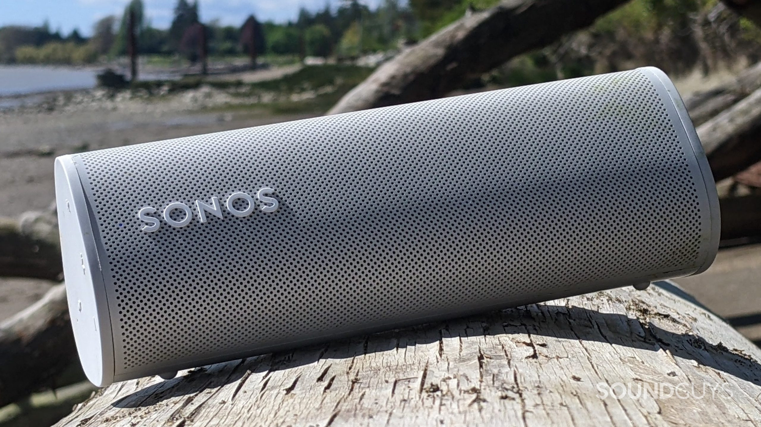 sonos-roam-outdoors-3 A white Sonos Roam speaker sitting at a slight angle on a log on a beach in front of a fallen tree and a body of water along with some trees in the distance.