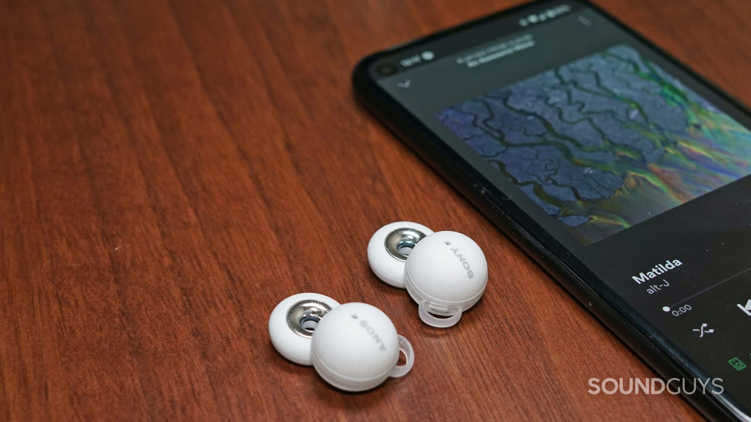 Sony-Linkbuds-phone-shot The Sony LinkBuds lay on a wooden surface next to a Google Pixel 4a running Spotify playing Matilda by alt-J.