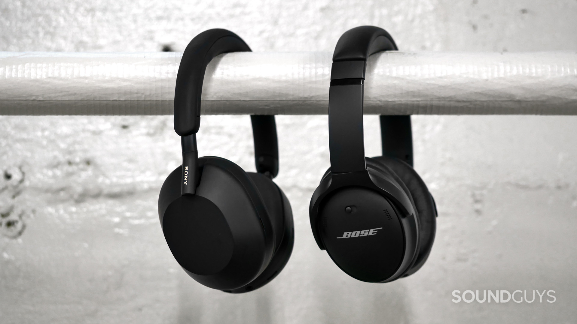 sony-wh-1000xm5-vs-bose-quietcomfort-45 Side-on view of Sony WH-1000XM5 beside the Bose Quietcomfort 45 hanging over a white horizontal pipe