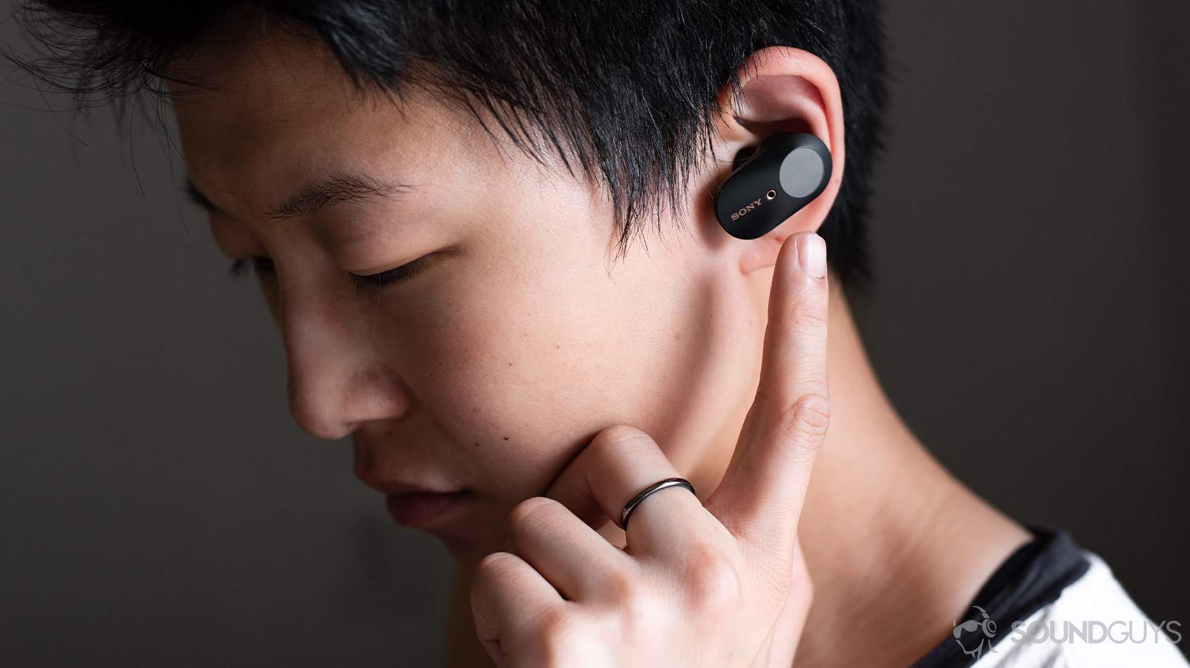 Sony WF-1000XM3-6_woman wearing earbud touch control gestures A photo of a woman wearing the Sony WF-1000XM3 earbuds and reaching for the touch panel of the left earbud. Meant to illustrate options for Sony headphones.