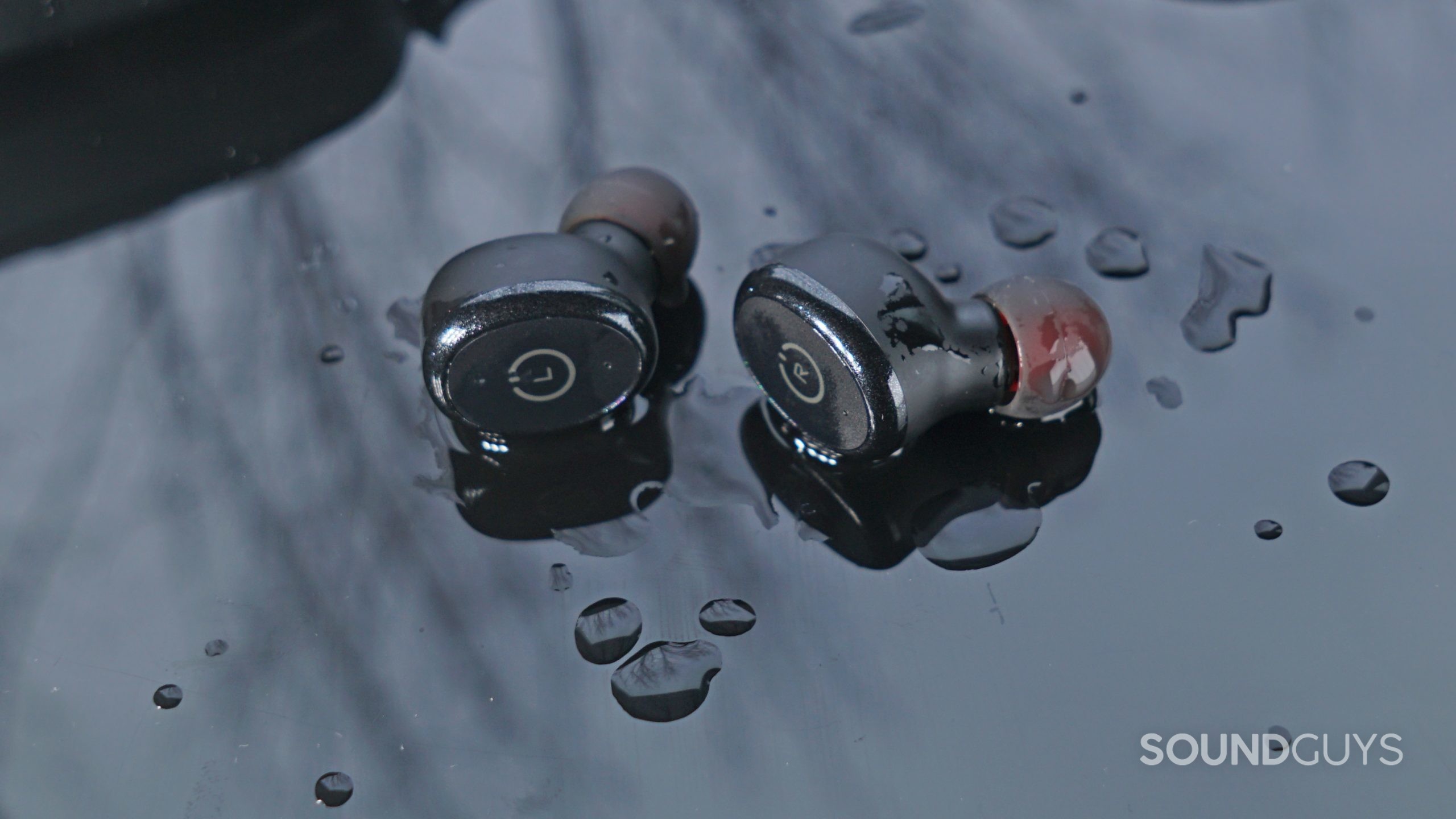 TOZO-T10-Wet-Buds The TOZO T10 earbuds lay on a reflective black surface covered in water.