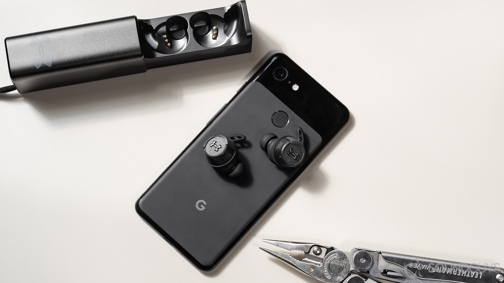 UA True Wireless FLASH X by JBL-workout earbuds buttons case Google Pixel 3 The UA True Wireless FLASH X by JBL on a Google Pixel 3 smartphone, next to the open charging case and a Leatherman Wave+ on a white surface.