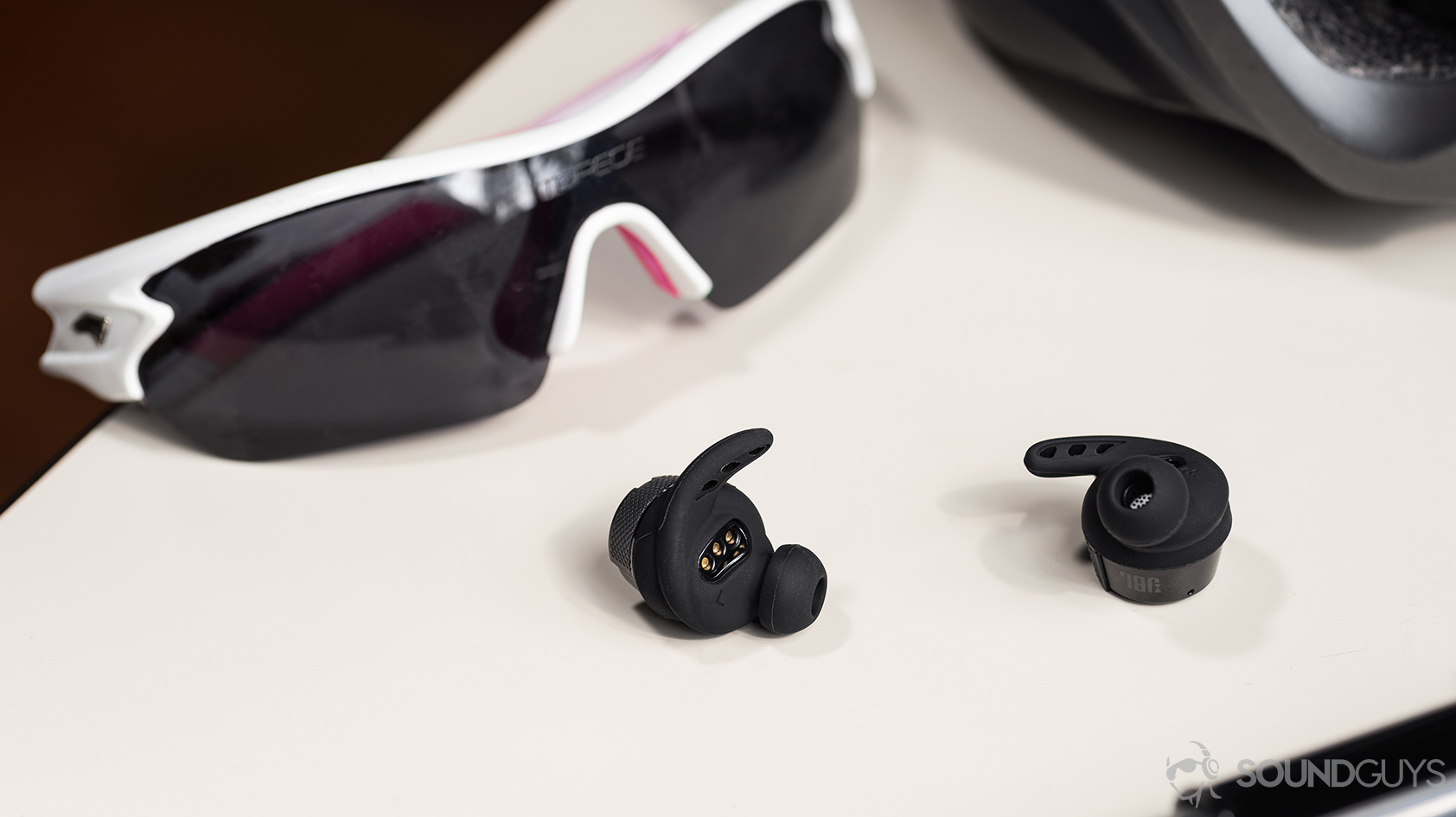 UA True Wireless FLASH X by JBL-workout earbuds wing ear tips biking glasses A photo of the UA True Wireless FLASH X by JBL with the wing and ear tips on display in front of cycling glasses on a white table.
